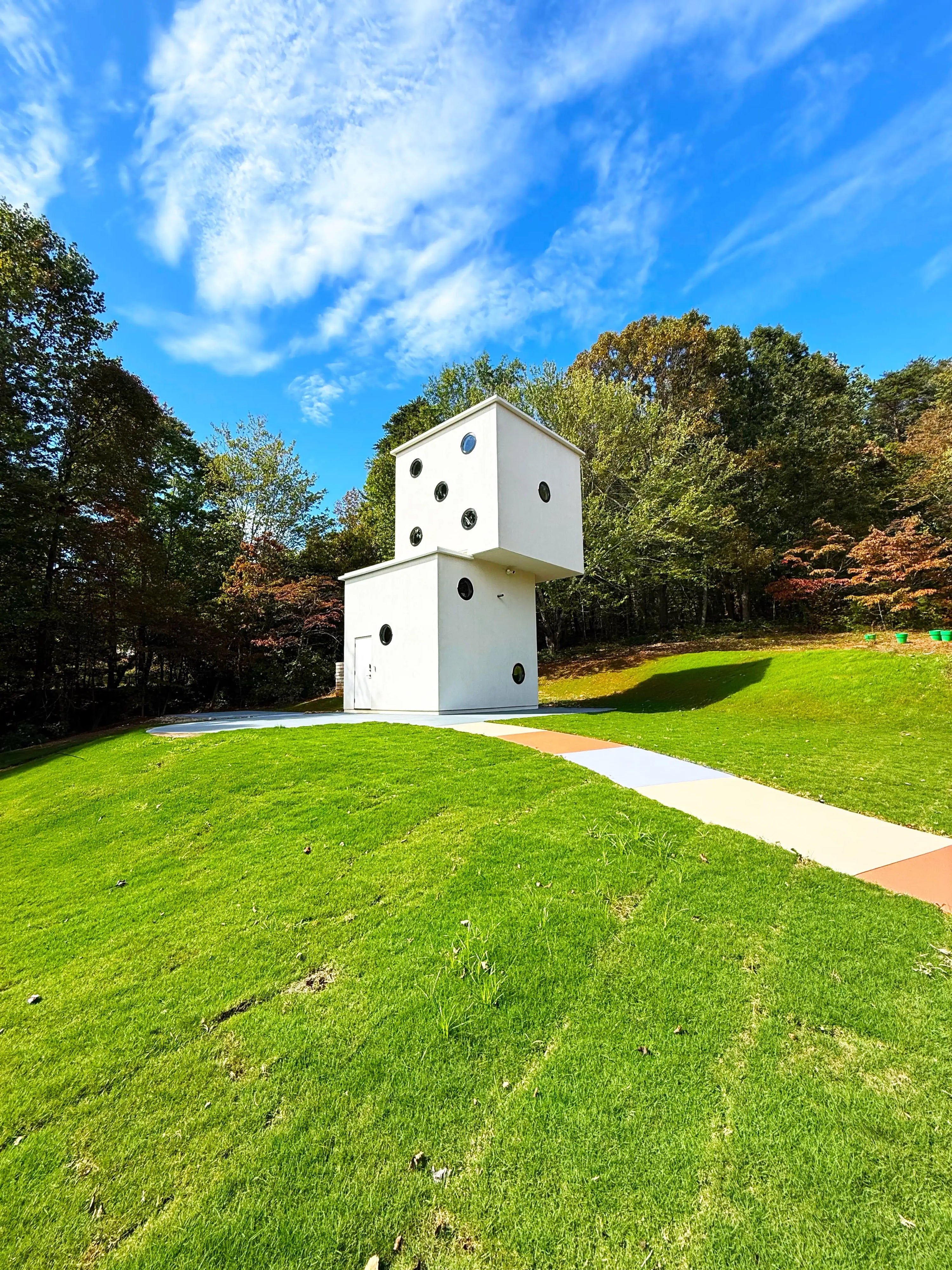 The Tiny Dice House, which is a tiny home in the shape of a pair of stacked dice is pictured atop a vibrantly green grassy hill, with a colorful pathway of alternating colors leading out from its foundation.
